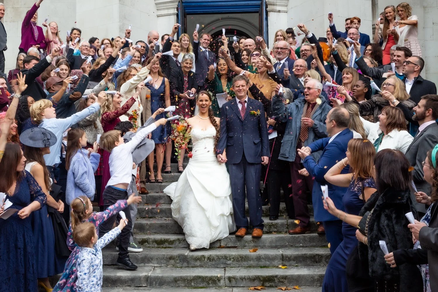 A bride and groom walking down the stairs surrounded by jubilant wedding guests throwing confetti and celebrating.