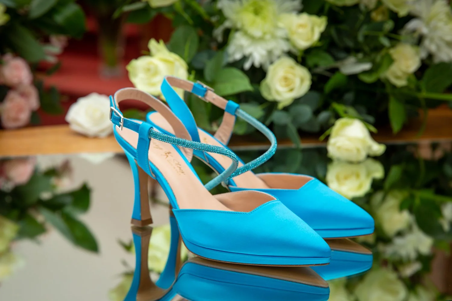 Blue high-heeled shoes with ankle straps and rhinestone embellishments, placed on a reflective surface in front of white roses.