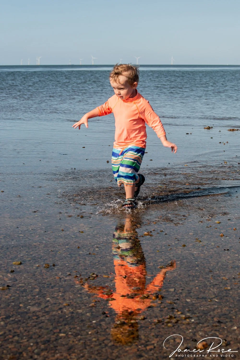 A young boy is playing in shallow water at the edge of a beach with wind turbines in the background.