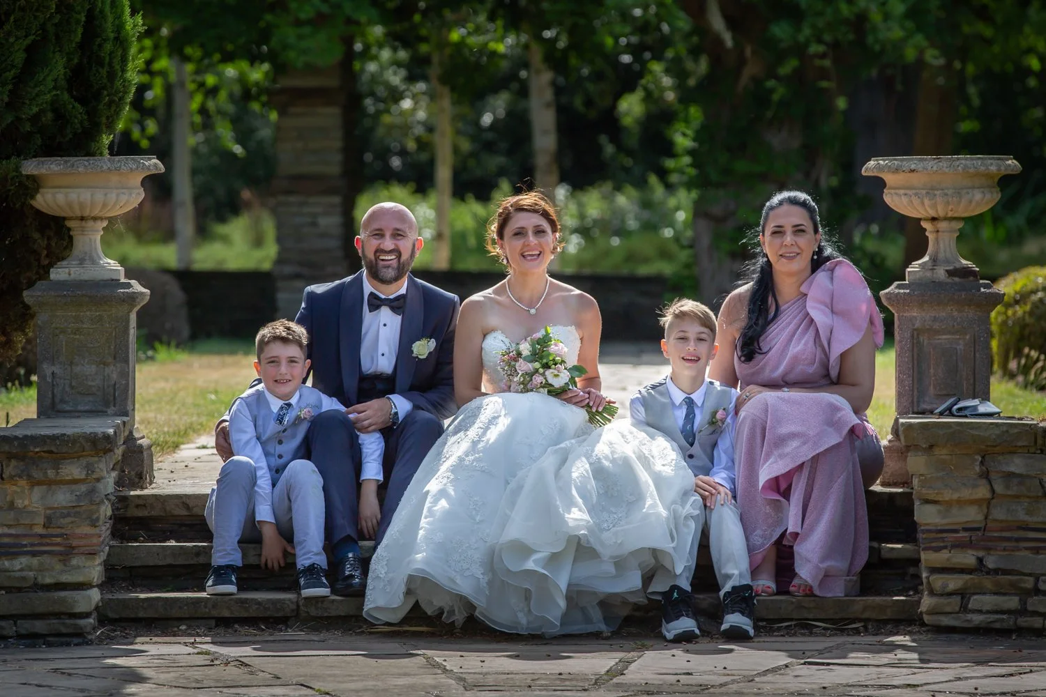 A wedding party sitting on stone steps outdoors, four adults and two children, with trees in the background. The bride in a white gown holding a bouquet, the groom in a tuxedo, a woman in a pink dress, and two young boys dressed in light suits.