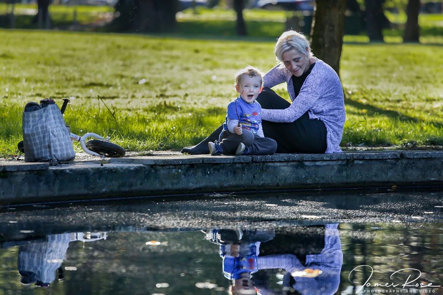 A woman and a young boy sitting on a park sidewalk near a pond, with a child's scooter and a bag nearby, reflected in the water.