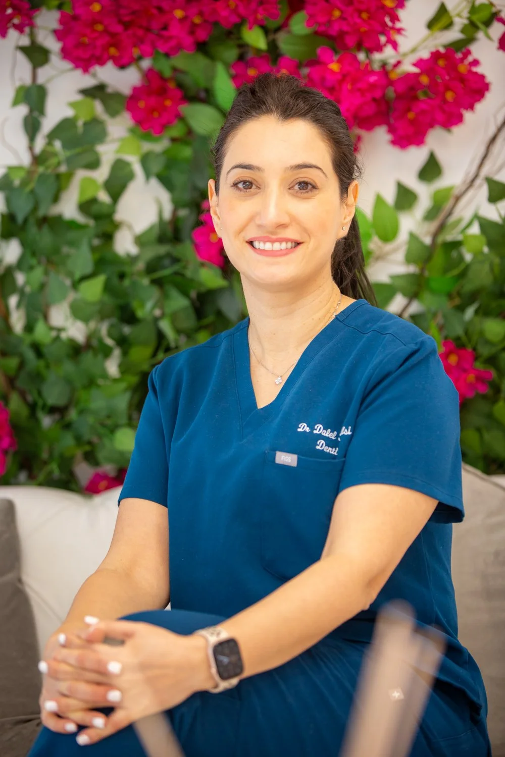 A smiling woman in blue medical scrubs sitting indoors with a background of green foliage and pink flowers.