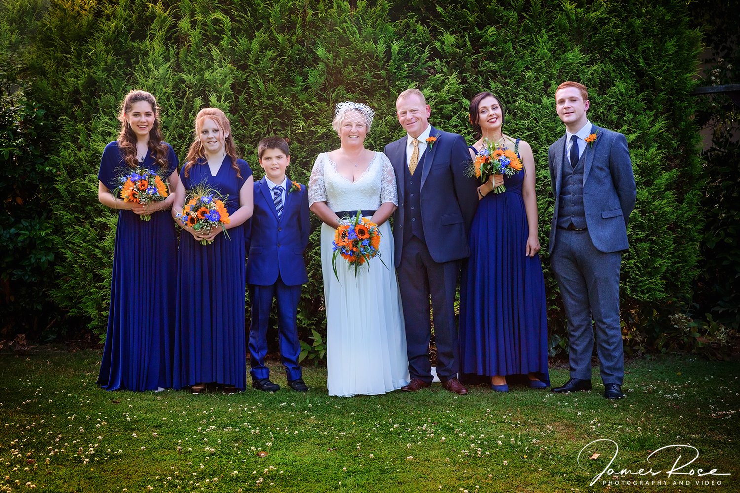 Group of wedding guests standing on grass with green bushes background, six women and one man dressed in formal attire, women holding bouquets of orange and blue flowers, smiling at camera.