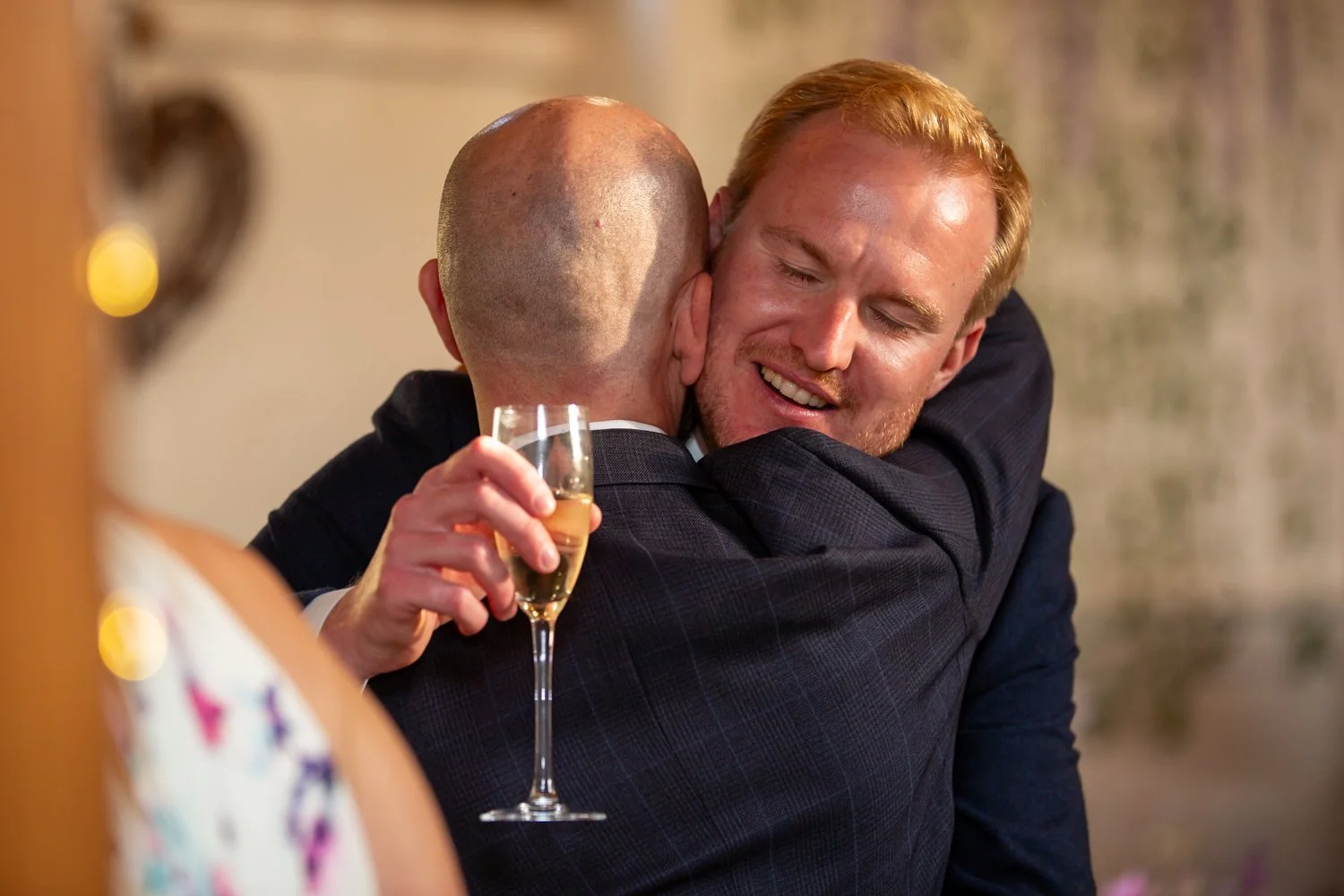 Two men hugging, one holding a glass of champagne, at a celebration or social gathering.