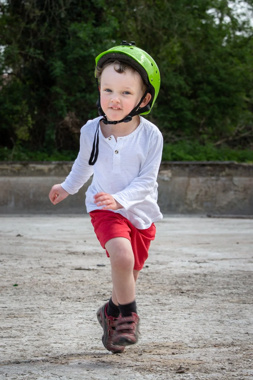 Young boy running outdoors wearing a green helmet, white long-sleeve shirt, red shorts, and athletic shoes, with trees in the background.