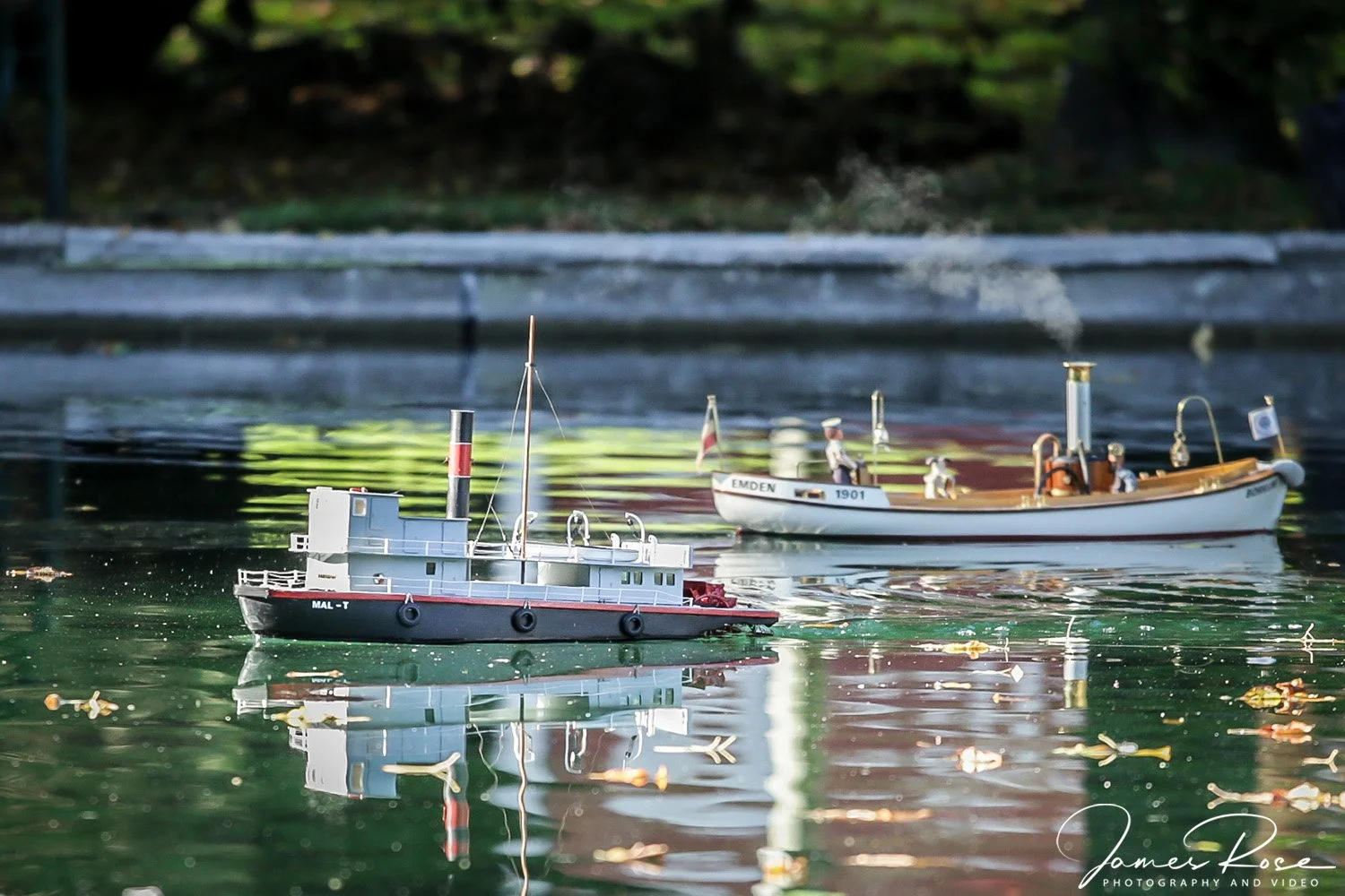 Model boats on a calm body of water, with one boat labeled 'MAL-T' and another labeled 'EMDEN 1901', surrounded by floating leaves and reflections.