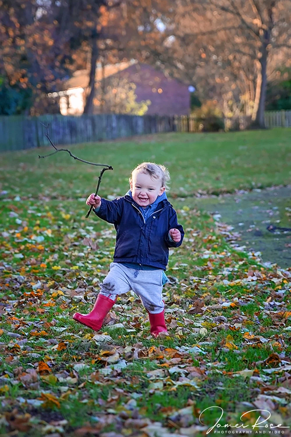 A young boy with blonde hair playing in a park on a fall day, holding a stick shaped like a small tree branch, with fallen leaves on the ground and a background of trees and a house.
