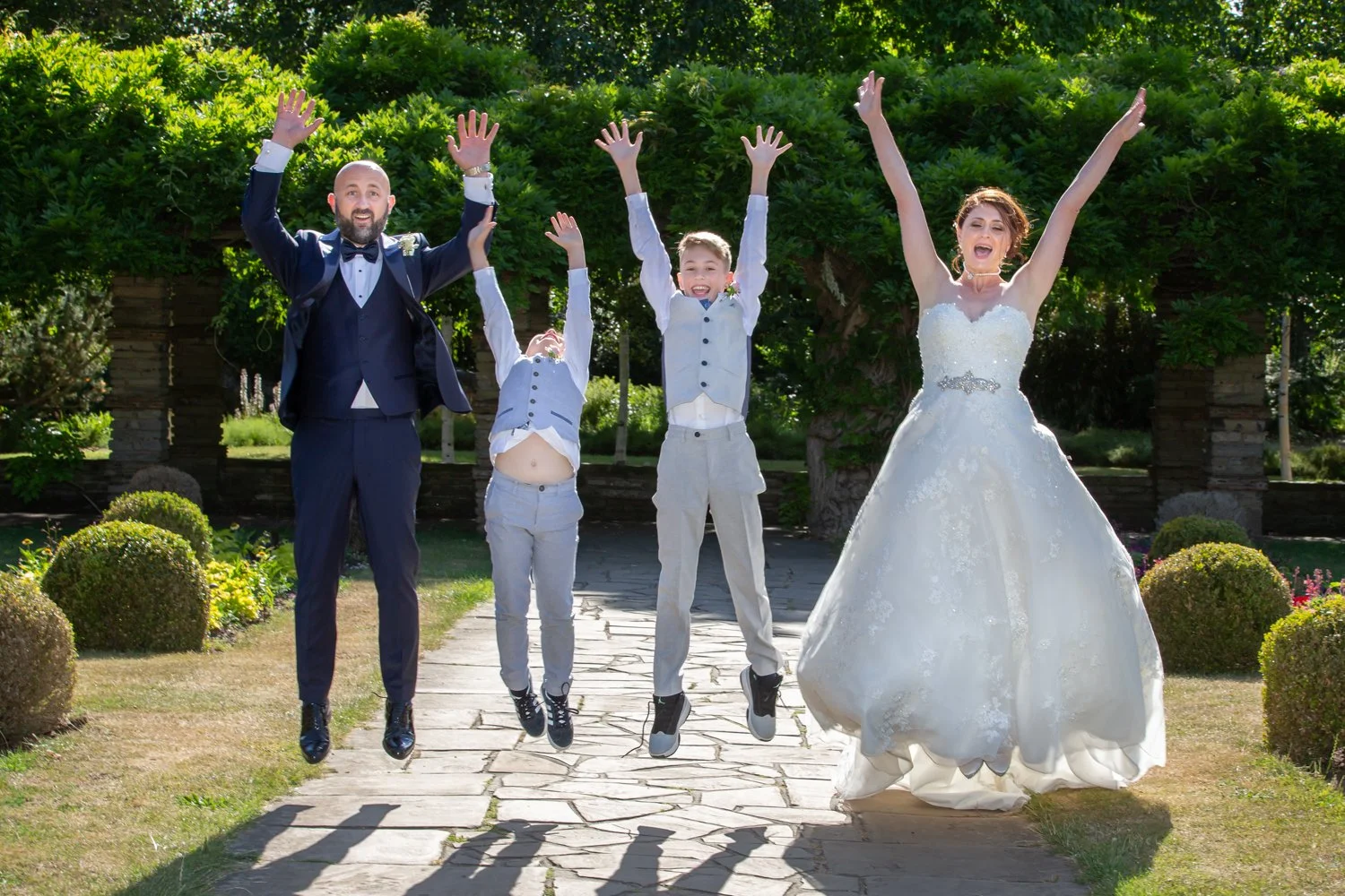 A wedding celebration in an outdoor garden with a man in a tuxedo, a woman in a wedding gown, and two young boys in formal attire, all jumping with their arms raised.