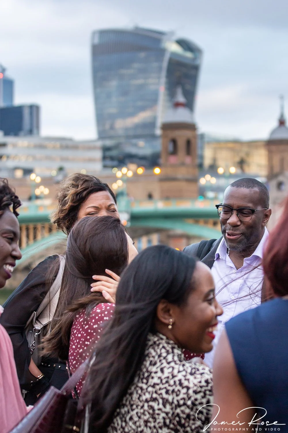 Group of diverse people smiling and hugging outdoors with City Hall and modern skyscrapers in London in the background.