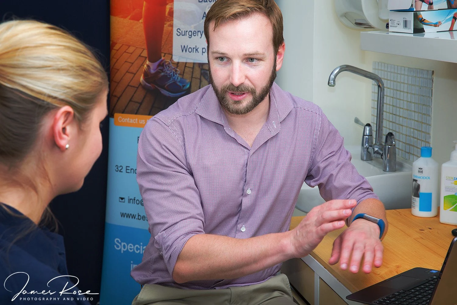 A man sitting at a table with a laptop, wearing a smartwatch and talking to a woman, in a medical or clinical setting, with bottles of sanitizer and medical equipment in the background.