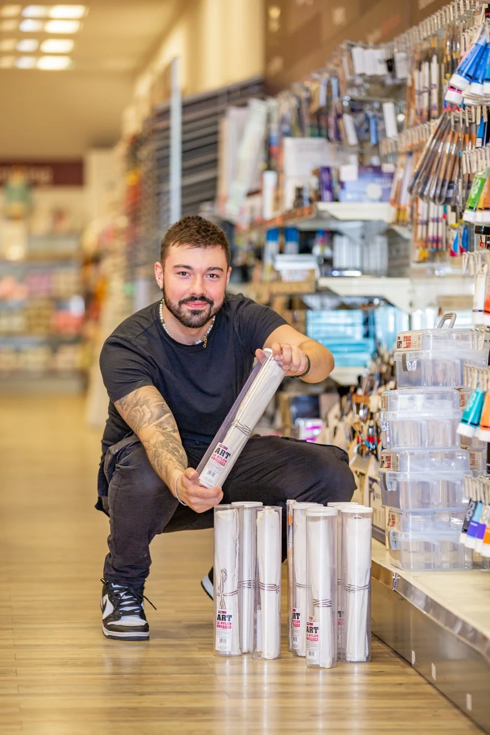 A man with tattoos and a beard crouching in an art supply store, holding a bundle of white canvas rolls in front of a shelf filled with art supplies.