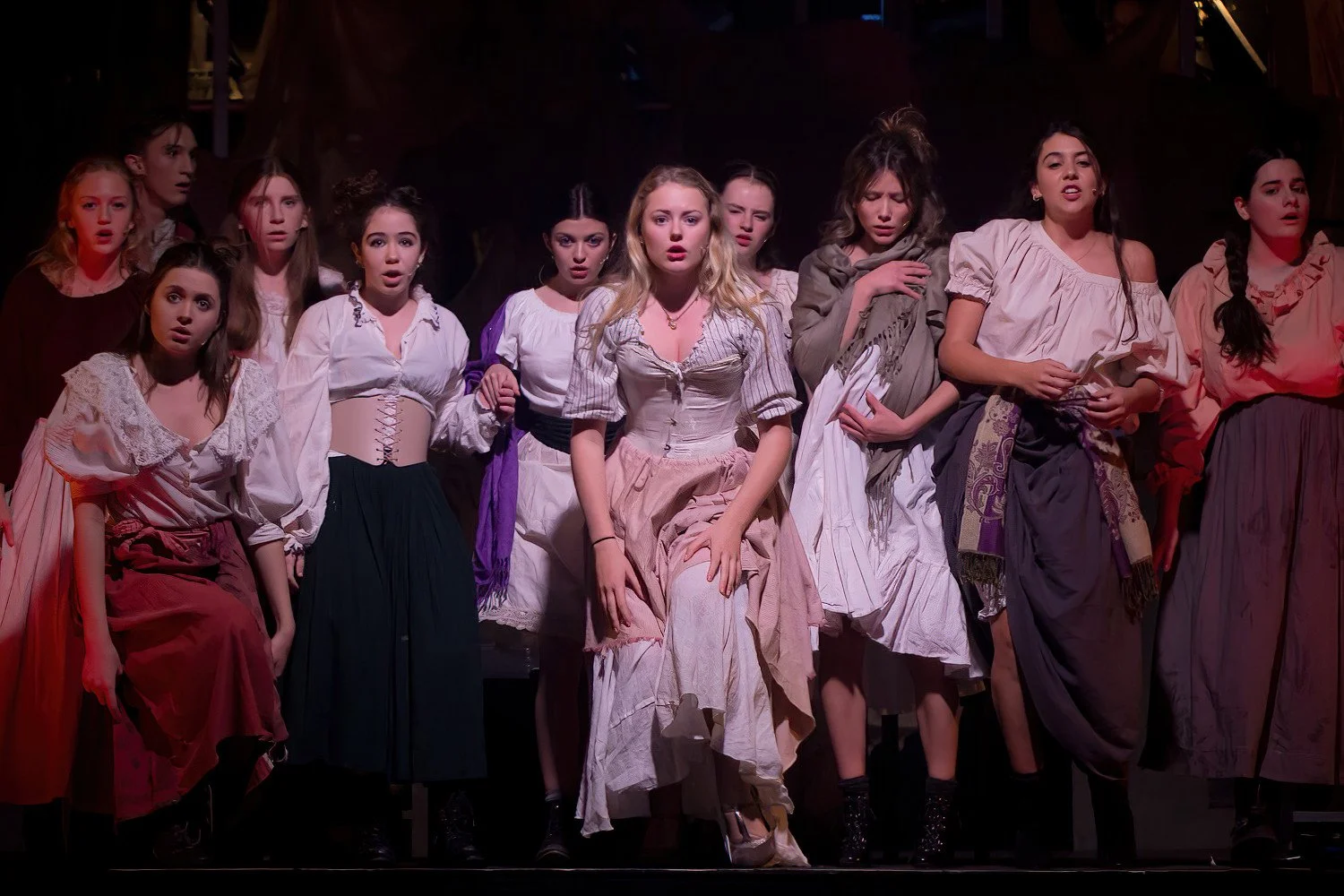 A group of young women dressed in period costumes on stage, appearing surprised or emotional during a theatrical performance.