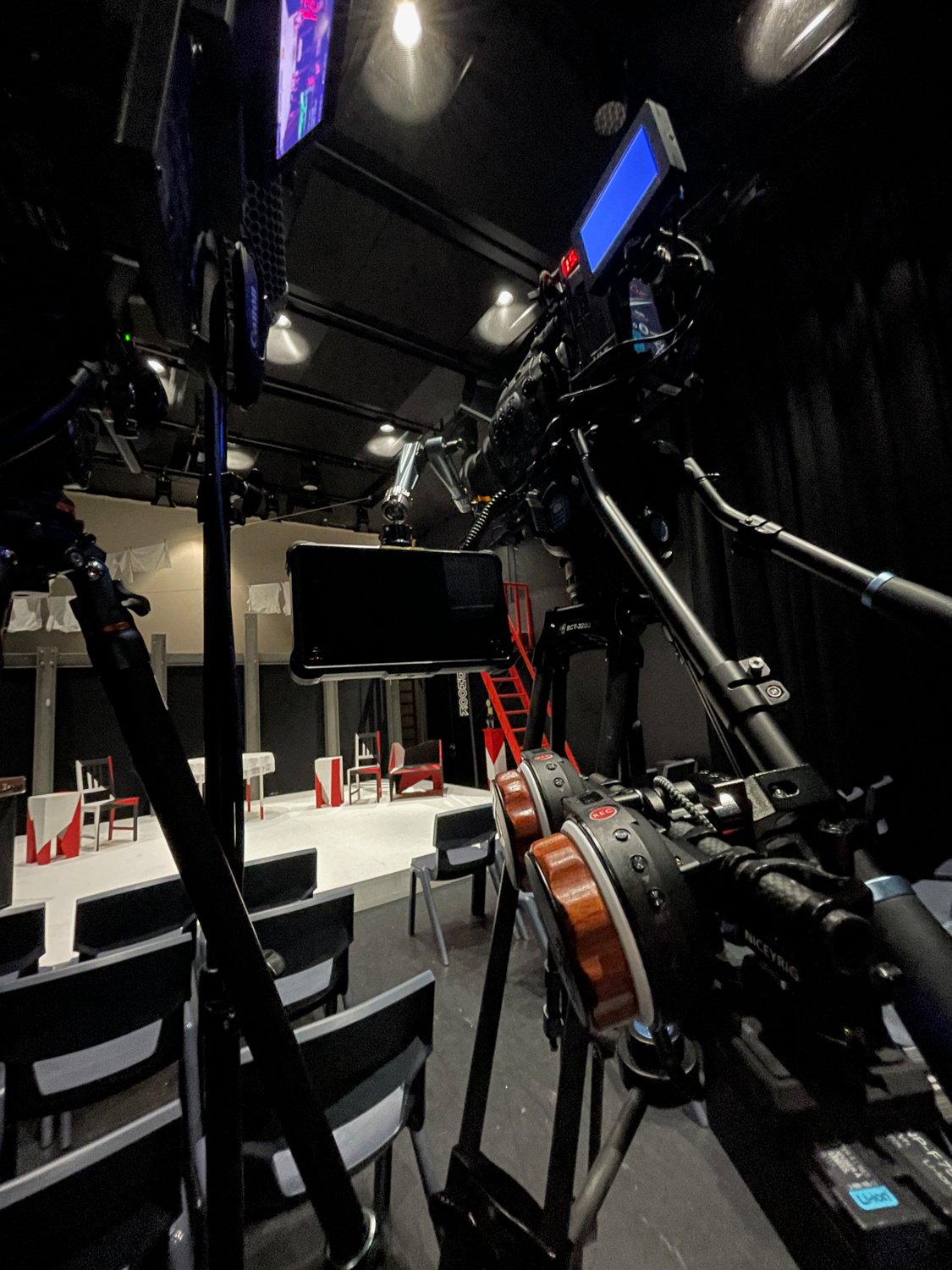 The image shows a film or television set with cameras and equipment pointed towards a small stage set with chairs and tables, prepared for a performance or interview.