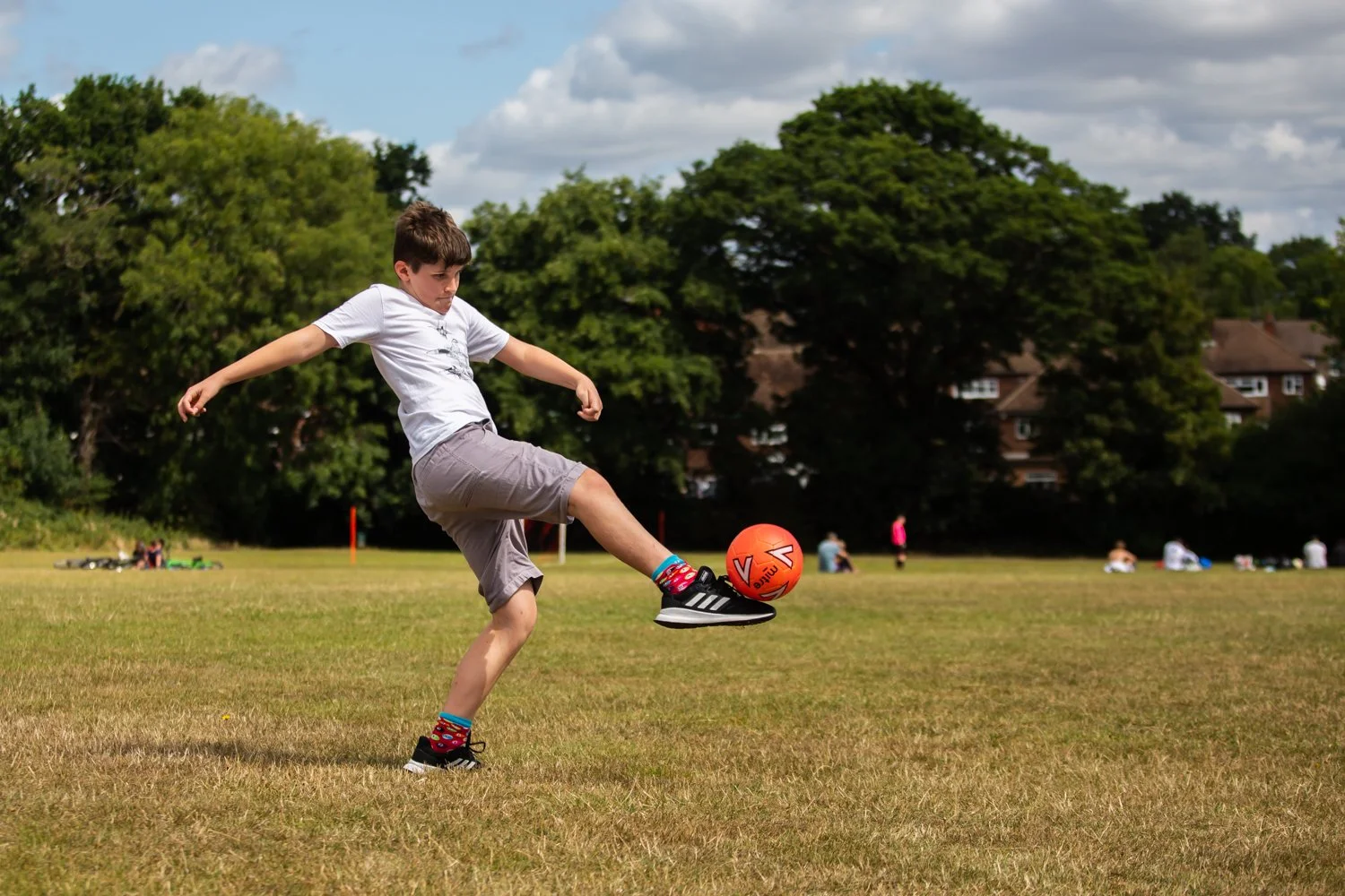 A boy playing soccer on a grassy field, kicking an orange ball with his right foot. He's wearing a white t-shirt, gray shorts, black sneakers, and colorful socks. There are trees and people in the background, with houses and a cloudy sky above.
