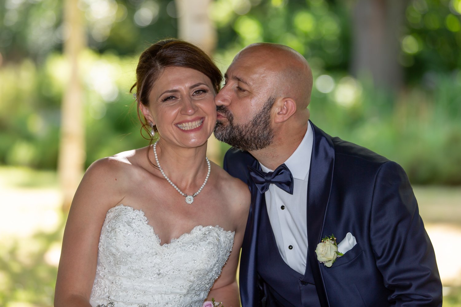 A bride and groom at their wedding; the groom is kissing the bride on the cheek, and she is smiling happily.