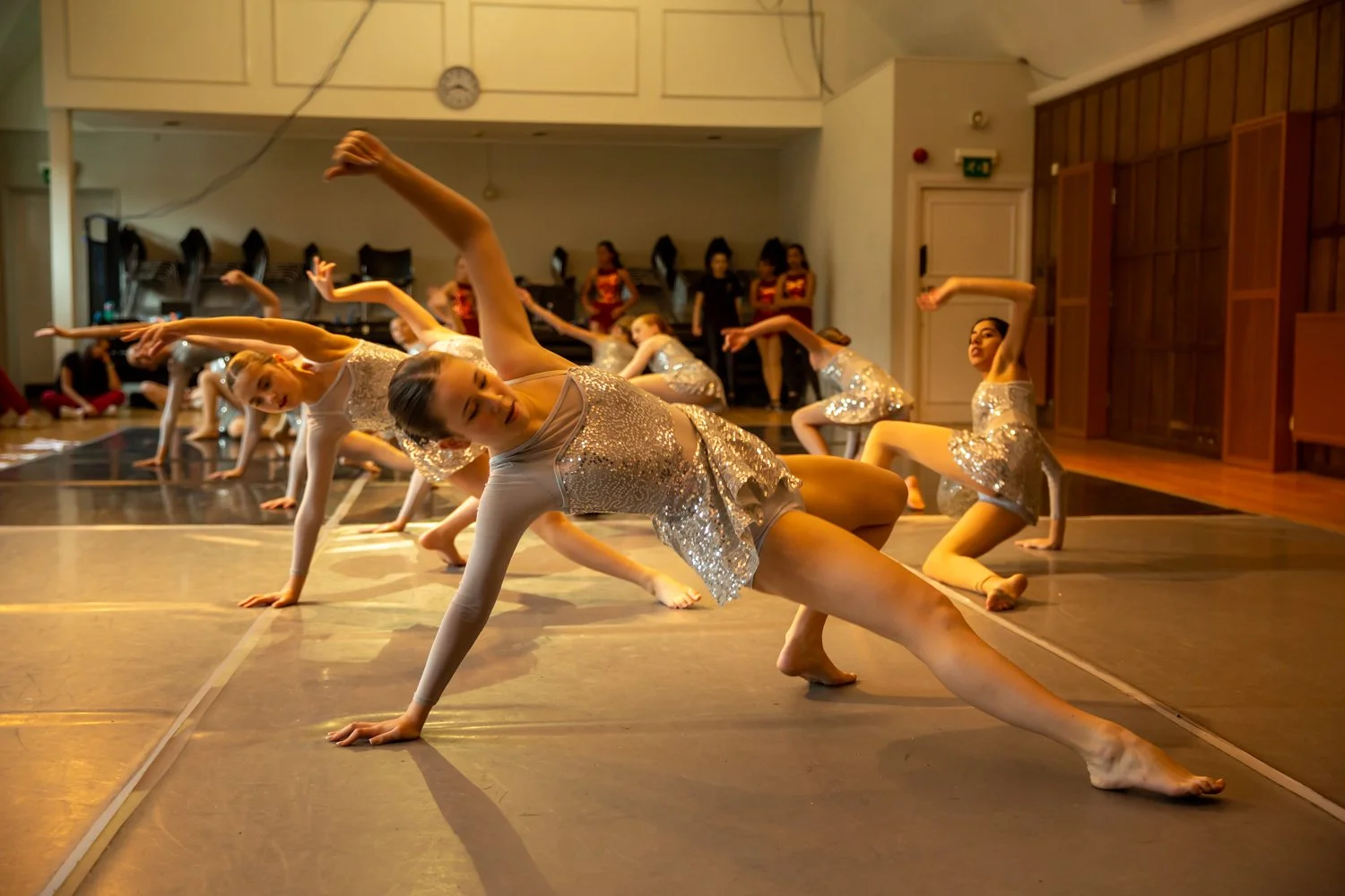 Young girls in shiny silver dance costumes performing a synchronized dance routine in a gym or dance studio.