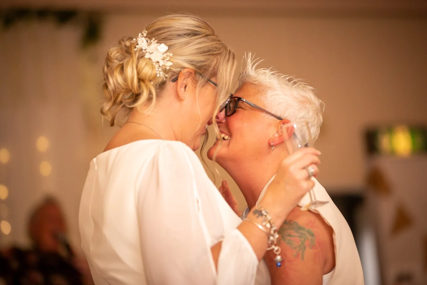 Two women with short, spiky gray hair and glasses smiling and touching foreheads at a celebration, one holding a glass of drink.