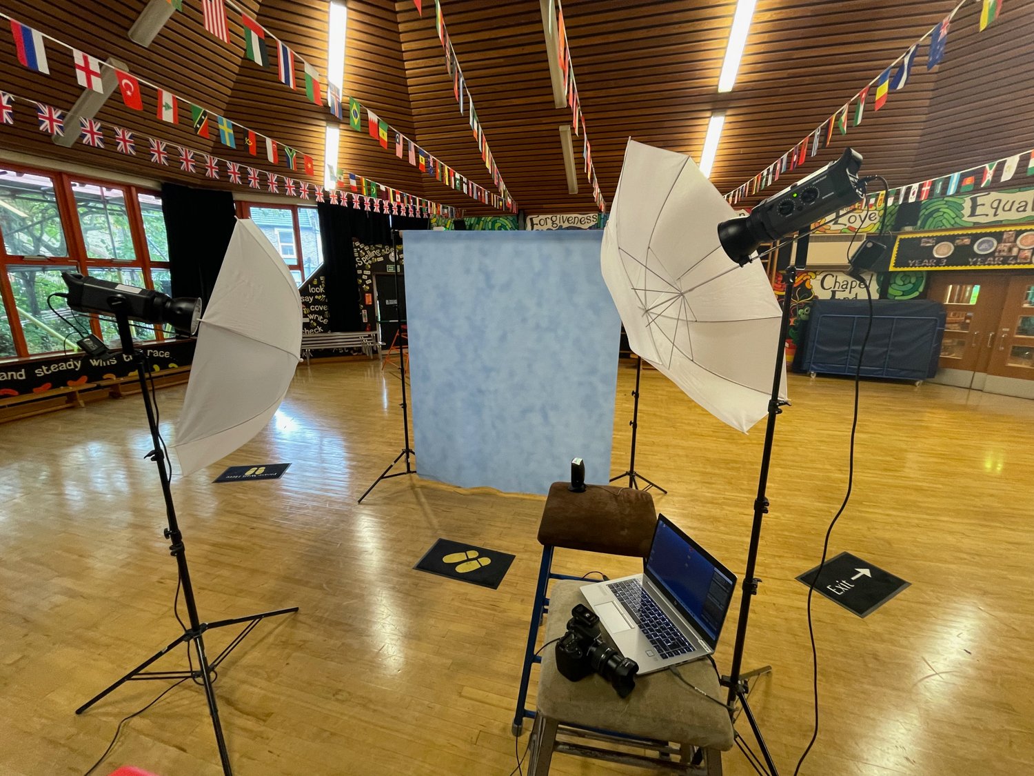 Set up for a photo shoot or video recording with two white umbrellas, a blue backdrop, a laptop, a camera, and lighting equipment in a spacious room with wooden floors and colorful flag banners hanging from the ceiling.