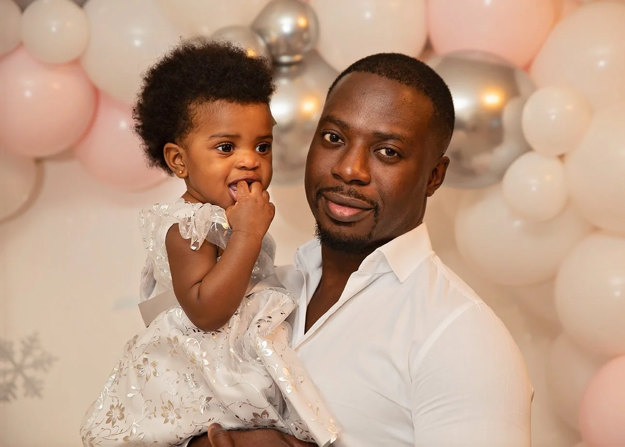 A man holding a young girl in front of a backdrop of pink, white, and silver balloons.