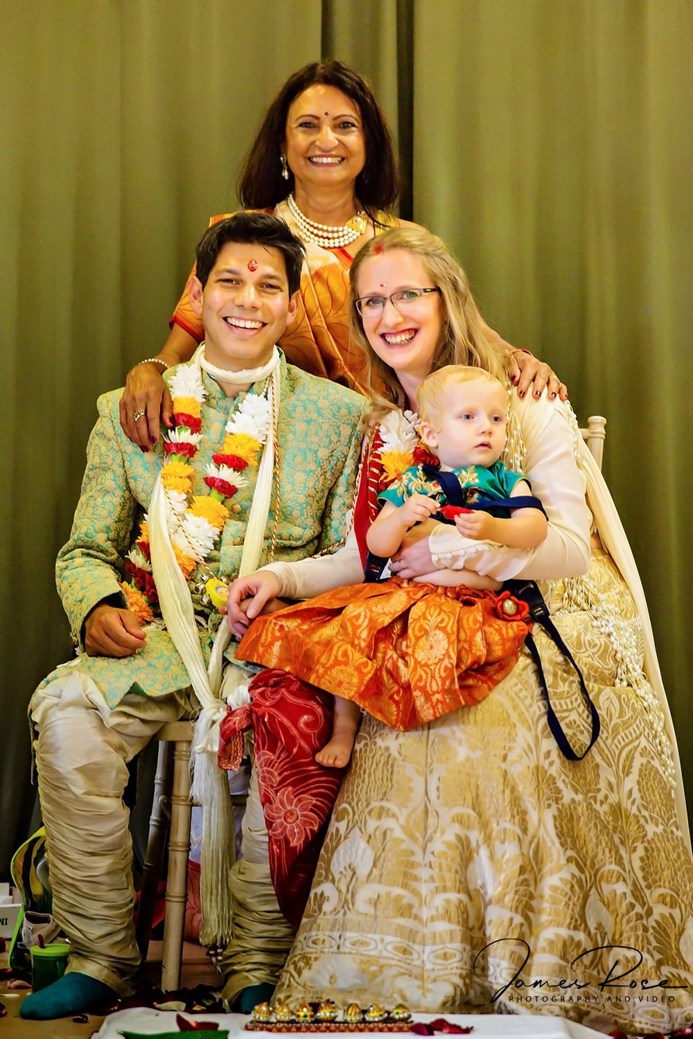 Group of four people at a traditional Indian wedding celebration, with the bride and groom in center, wearing traditional attire and flower garlands, posing with an older woman in an orange saree and two children, one of whom is sitting on the bride'