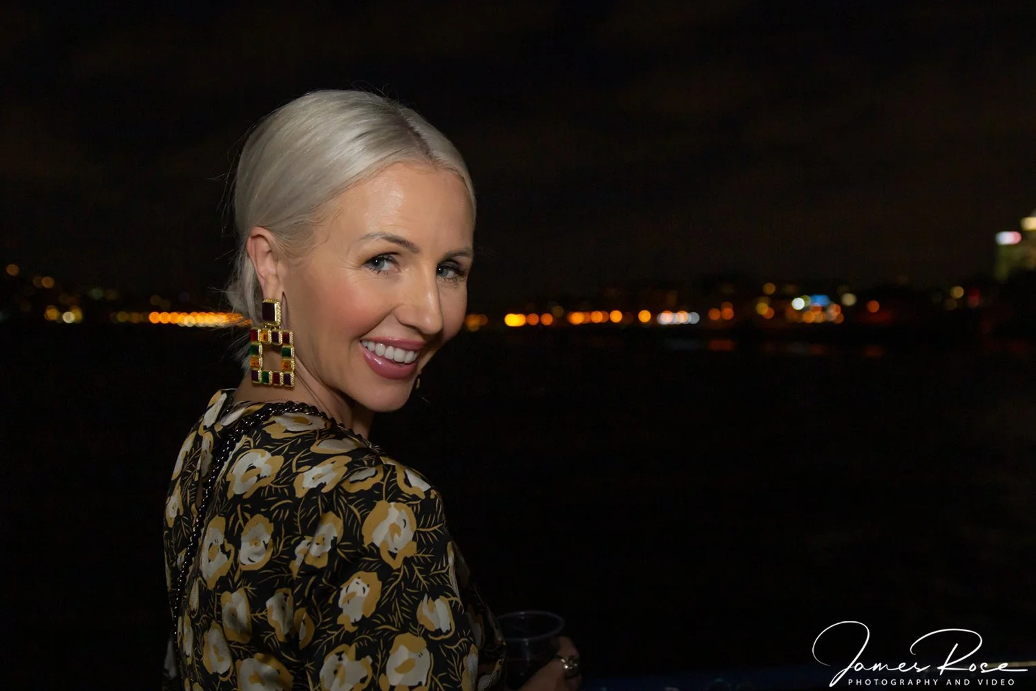 A smiling woman with platinum blonde hair at night, wearing large colorful earrings and a floral blouse, with city lights in the background.