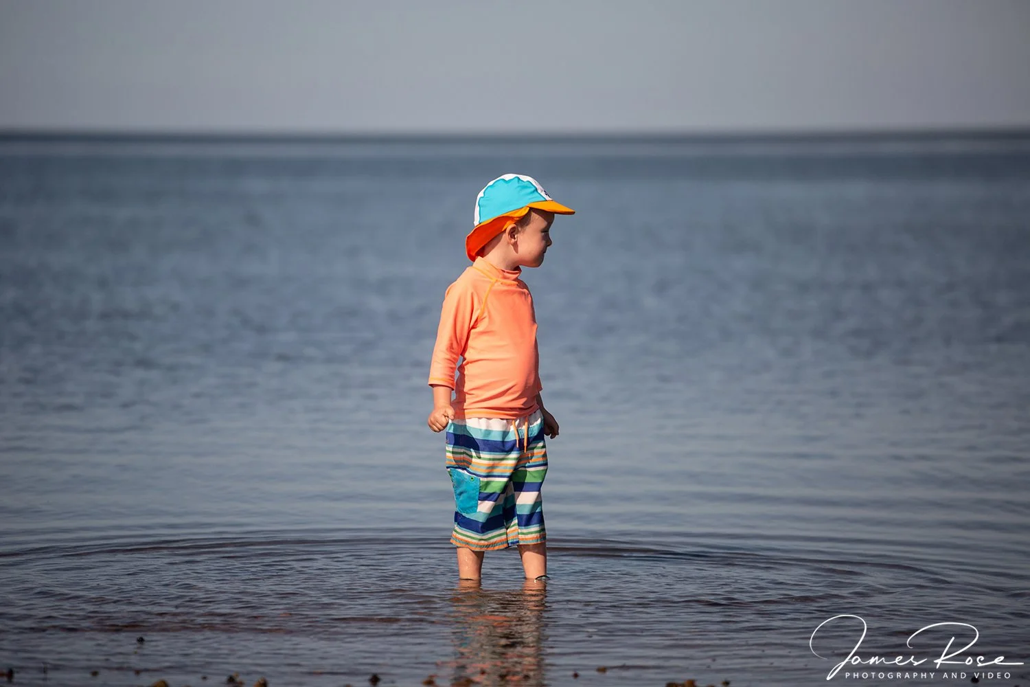 A young boy standing in shallow water at the beach, wearing colorful striped shorts, an orange long-sleeve shirt, and a multicolored cap, looking out over the water.