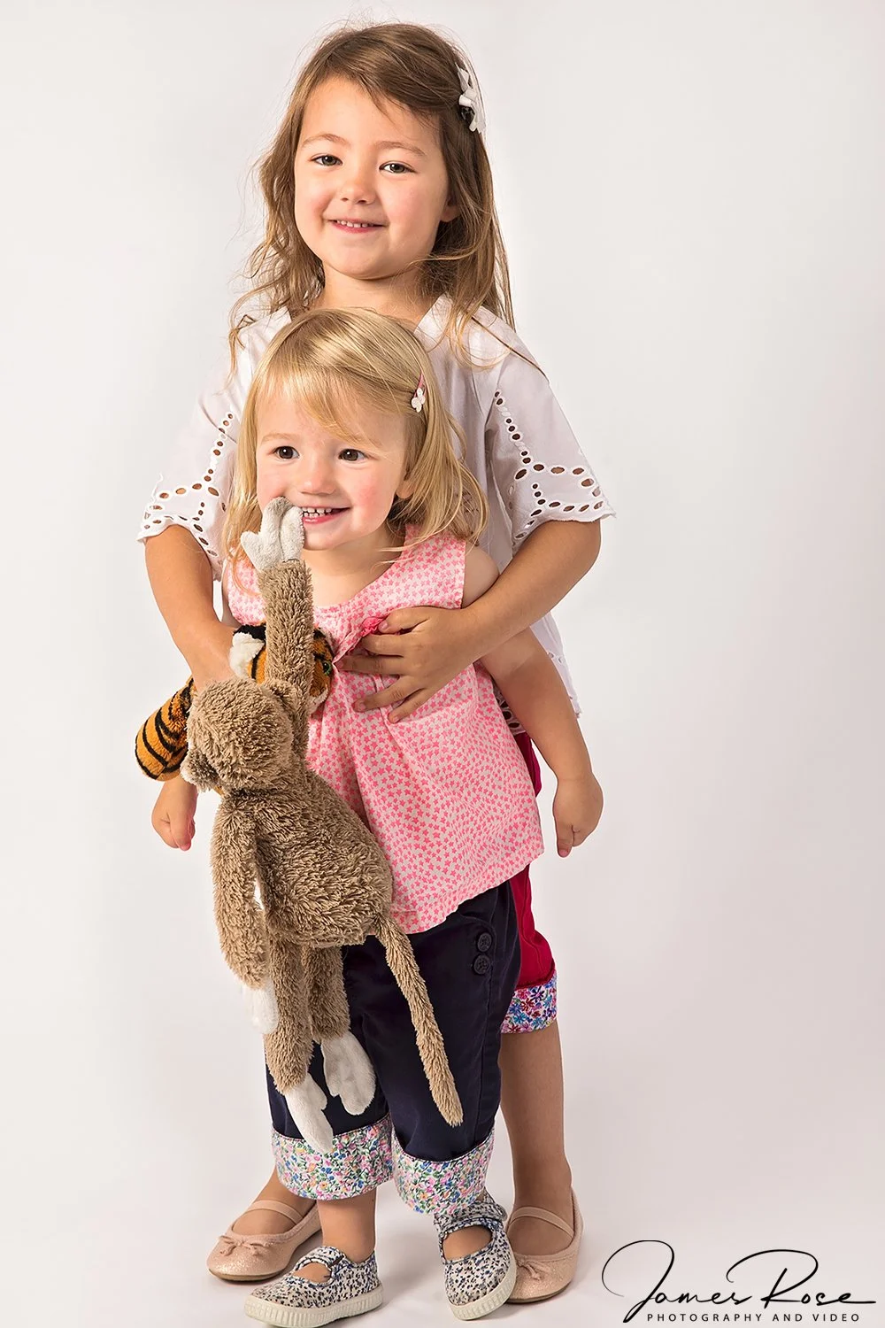 Two young girls smiling, one holding a plush animal and the other with her arms around her sister, in a studio setting.