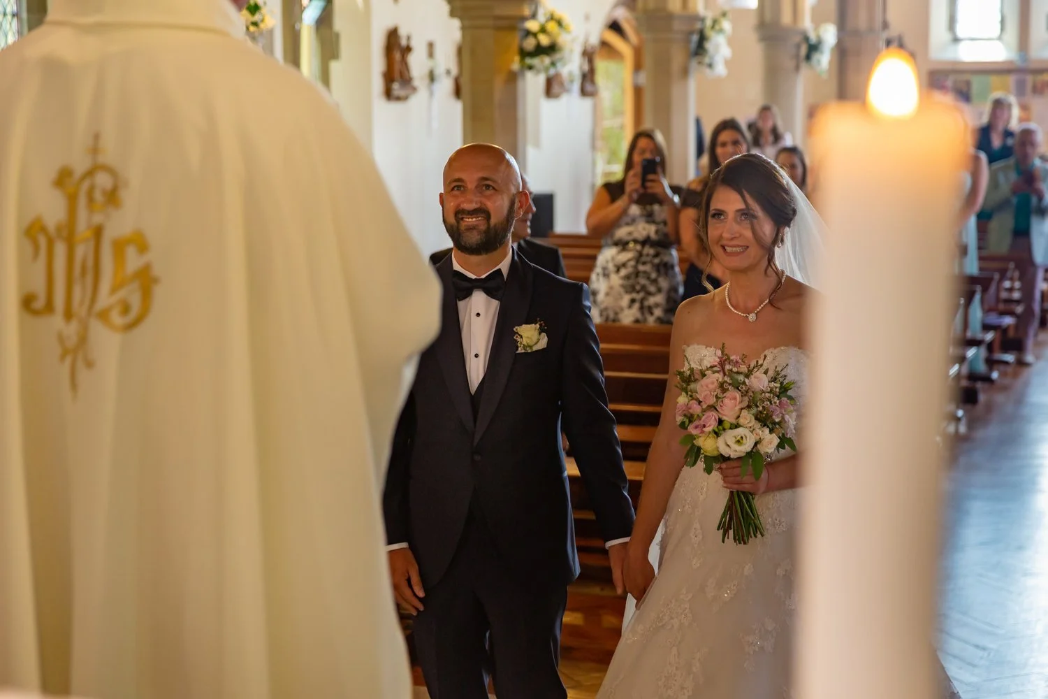 A bride and groom holding hands during their wedding ceremony inside a church, with guests seated in the background.