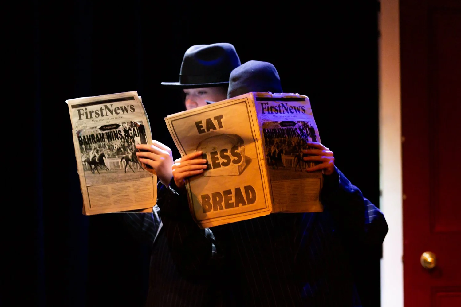 Two children dressed as newsroom reporters reading newspapers titled 'First News'. One child is wearing a fedora hat, the other a beanie, and they are standing under a dark background with a bold spotlight lighting their faces and newspapers.