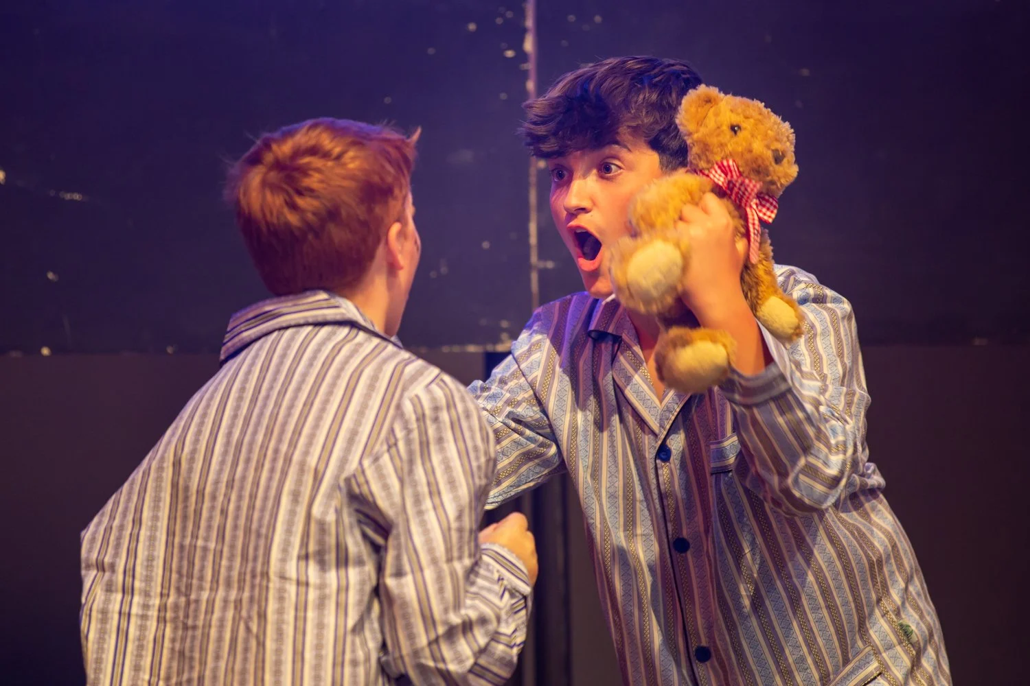 Two young males in striped pajamas having a heated argument on stage. One is holding a teddy bear with a surprised or angry expression.