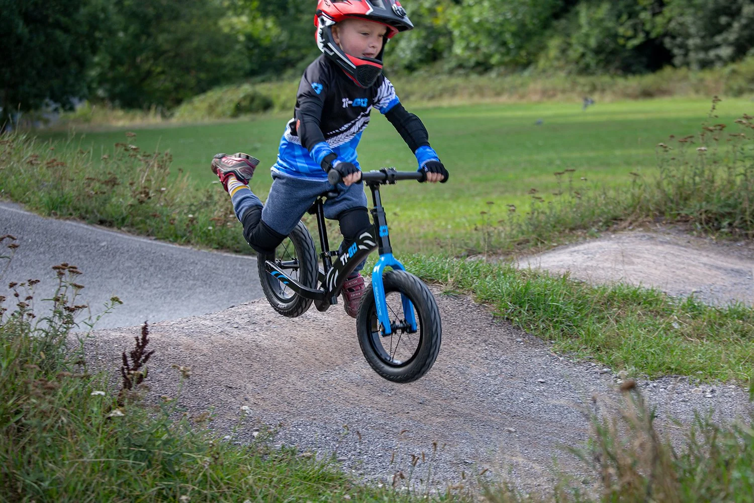 A young boy wearing a helmet and riding a small blue and black bike on a dirt track in a park surrounded by grass and trees.