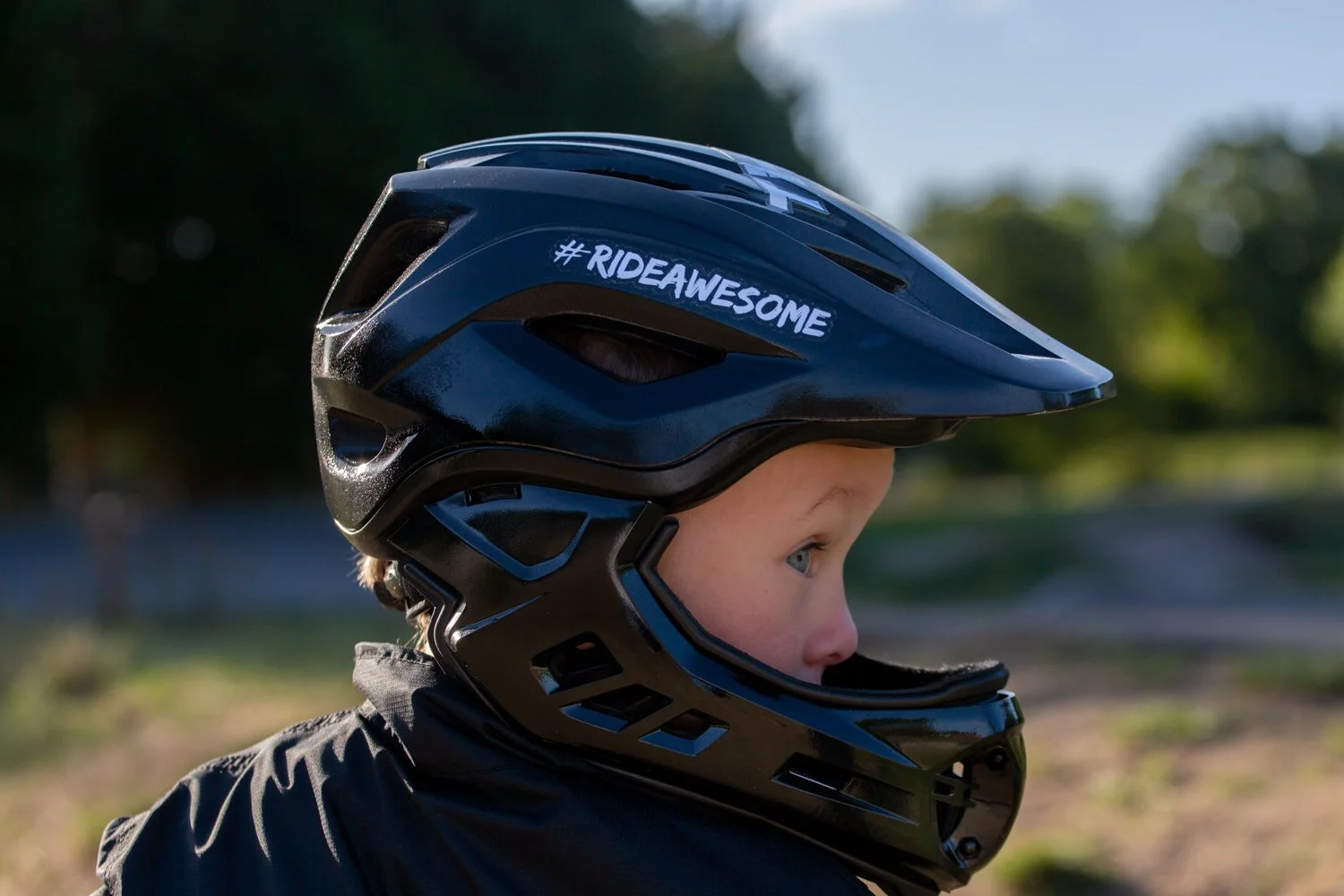 A young child wearing a black bicycle helmet outdoors with a hashtag #RIDEAWESOME written on it.