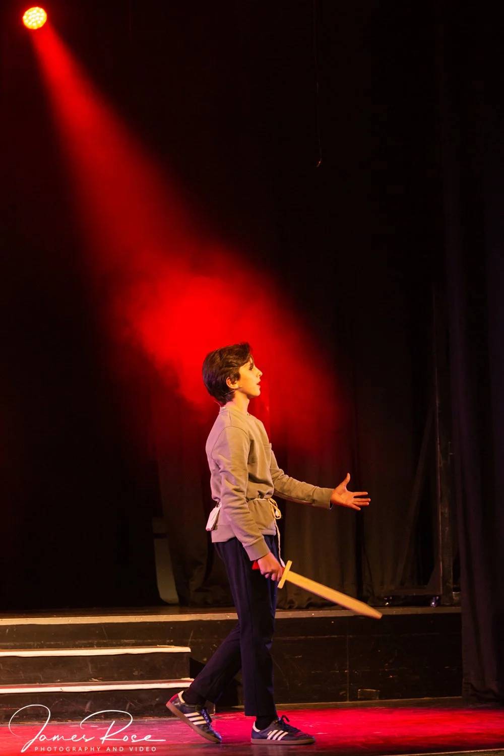 A young person dressed in casual attire, holding a wooden sword, appears to be performing or acting on stage under a red spotlight.