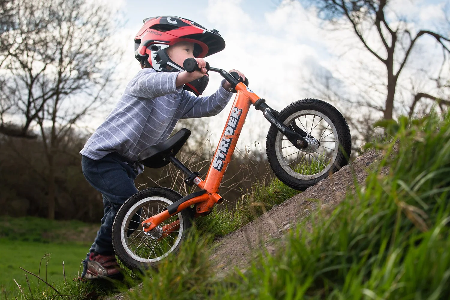 A young boy wearing a red and black helmet, gray striped shirt, and dark pants riding an orange balance bike uphill on a grassy slope outdoors with a cloudy sky and leafless trees in the background.