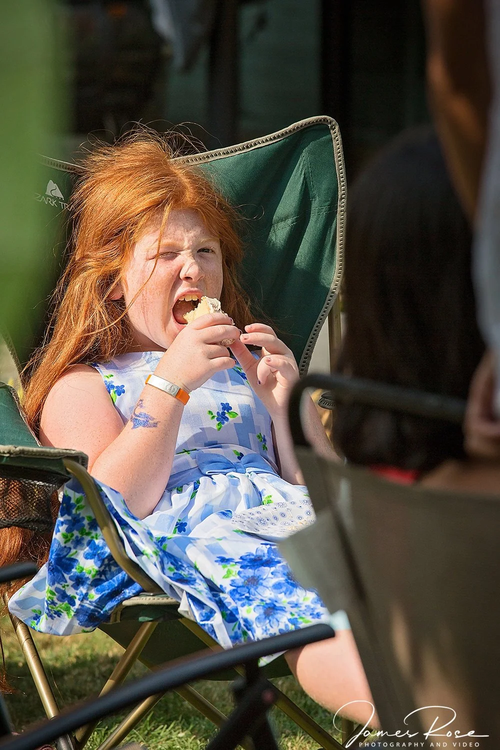A young girl with long red hair, sitting in a green outdoor chair, eating a piece of cake with one eye squinted closed.