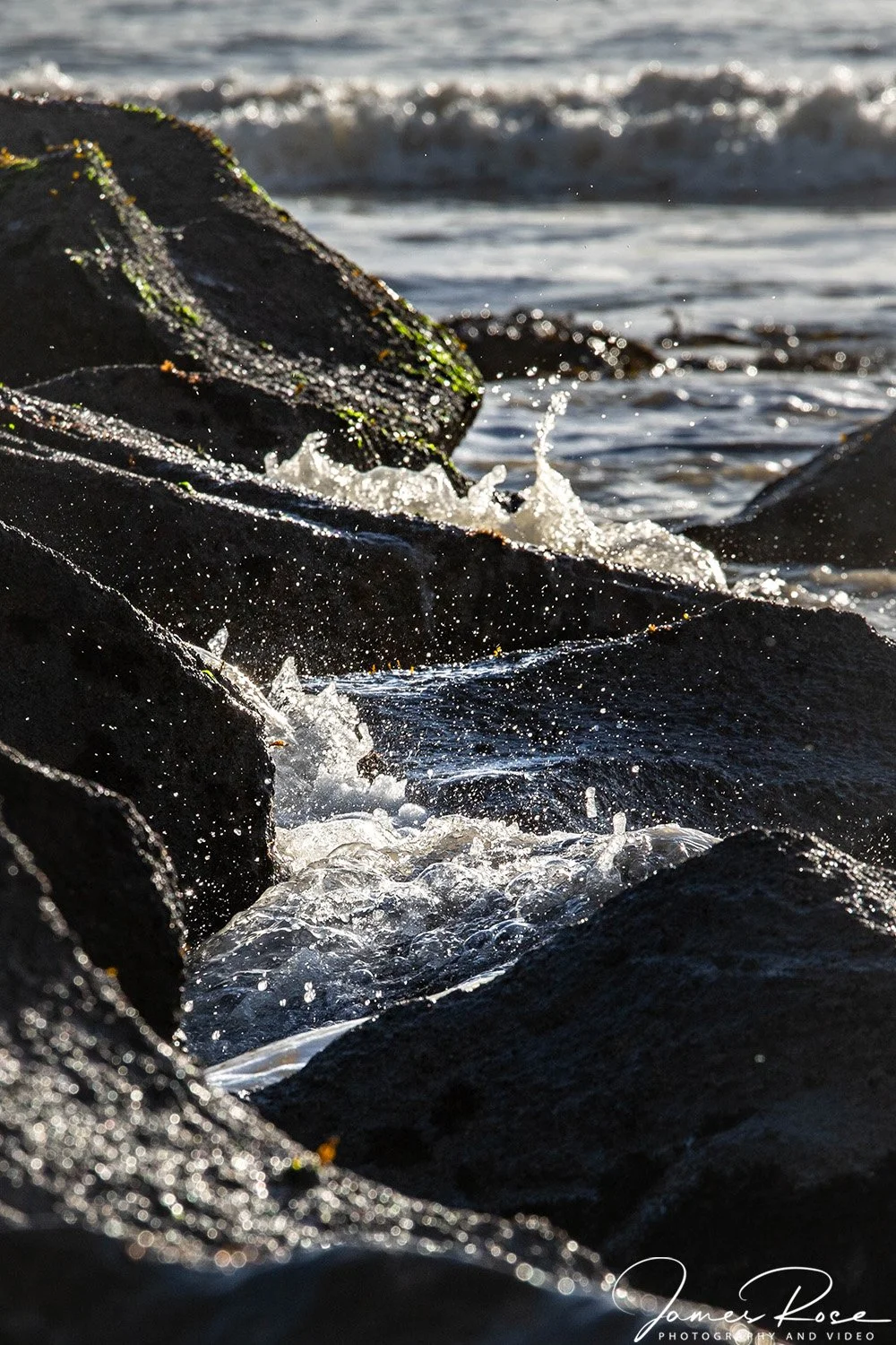 Waves crashing against black rocks on the shoreline with sunlight reflecting off the water.