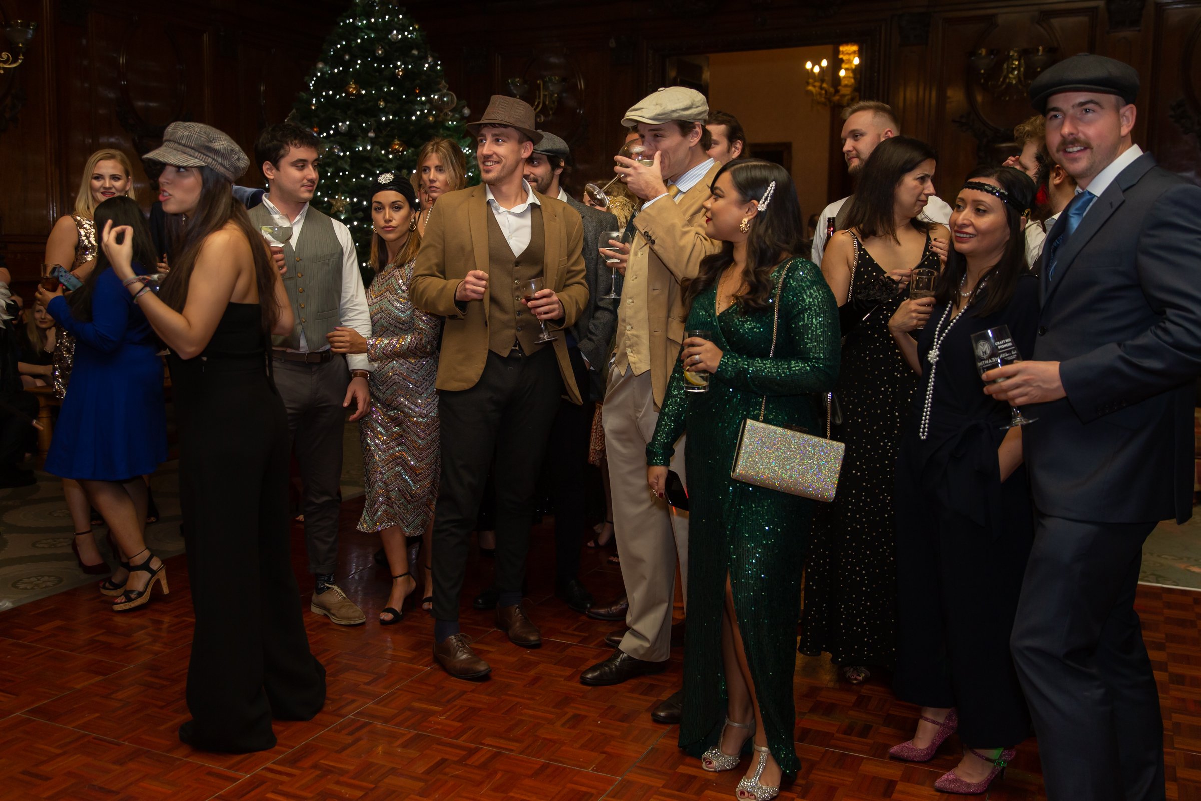 A group of people dressed in formal and vintage clothing at a holiday party with a decorated Christmas tree in the background.