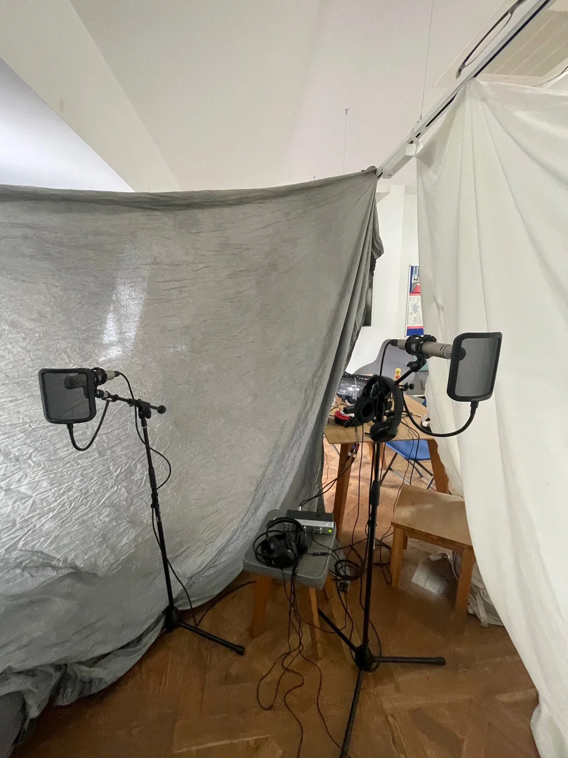 Recording studio setup with two microphones on stands, a pair of headphones, and a table with electronic equipment, all inside a room with white and gray curtains.