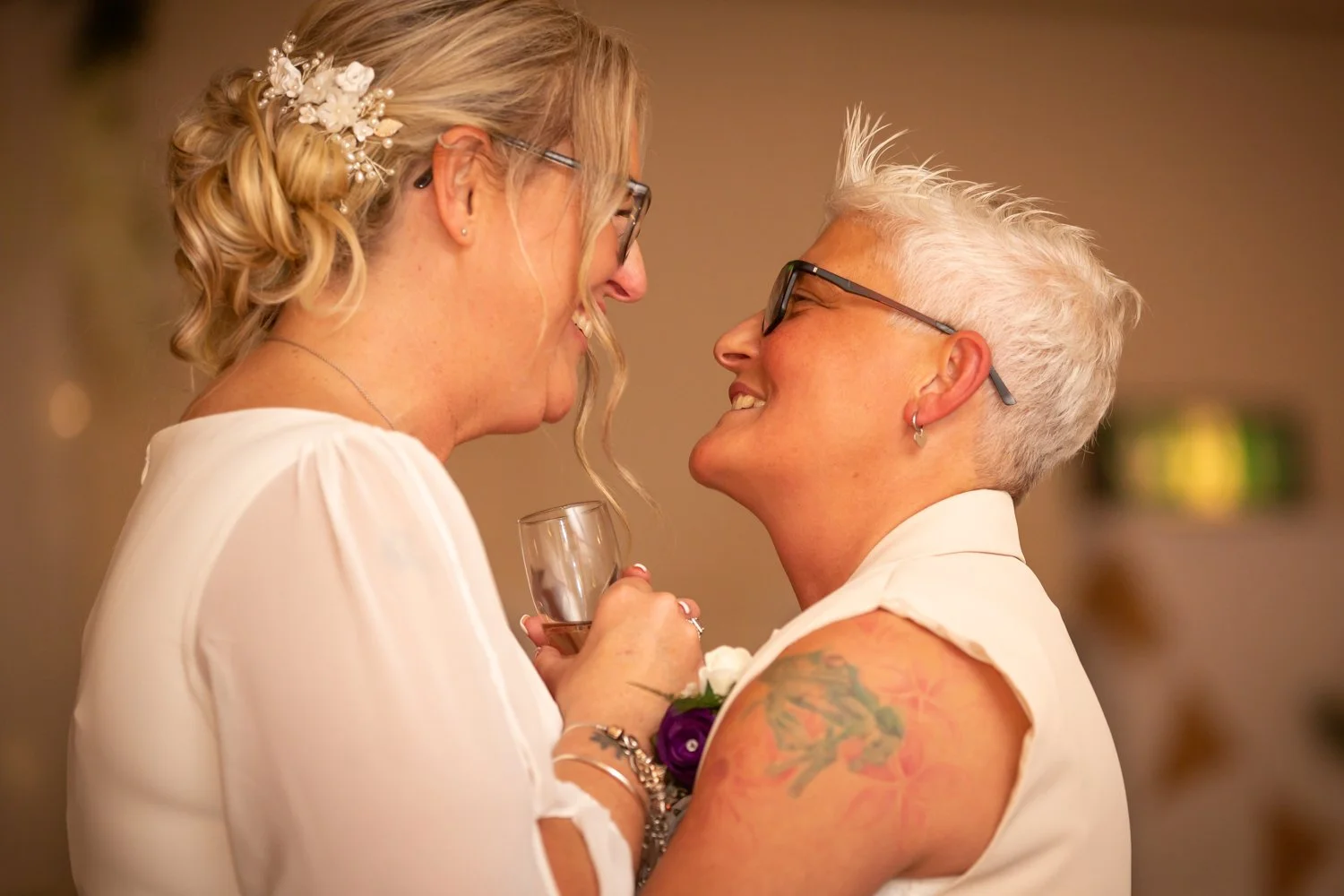 Two women celebrating together at a joyful event, one holding a glass of drink, both smiling and leaning close to each other.