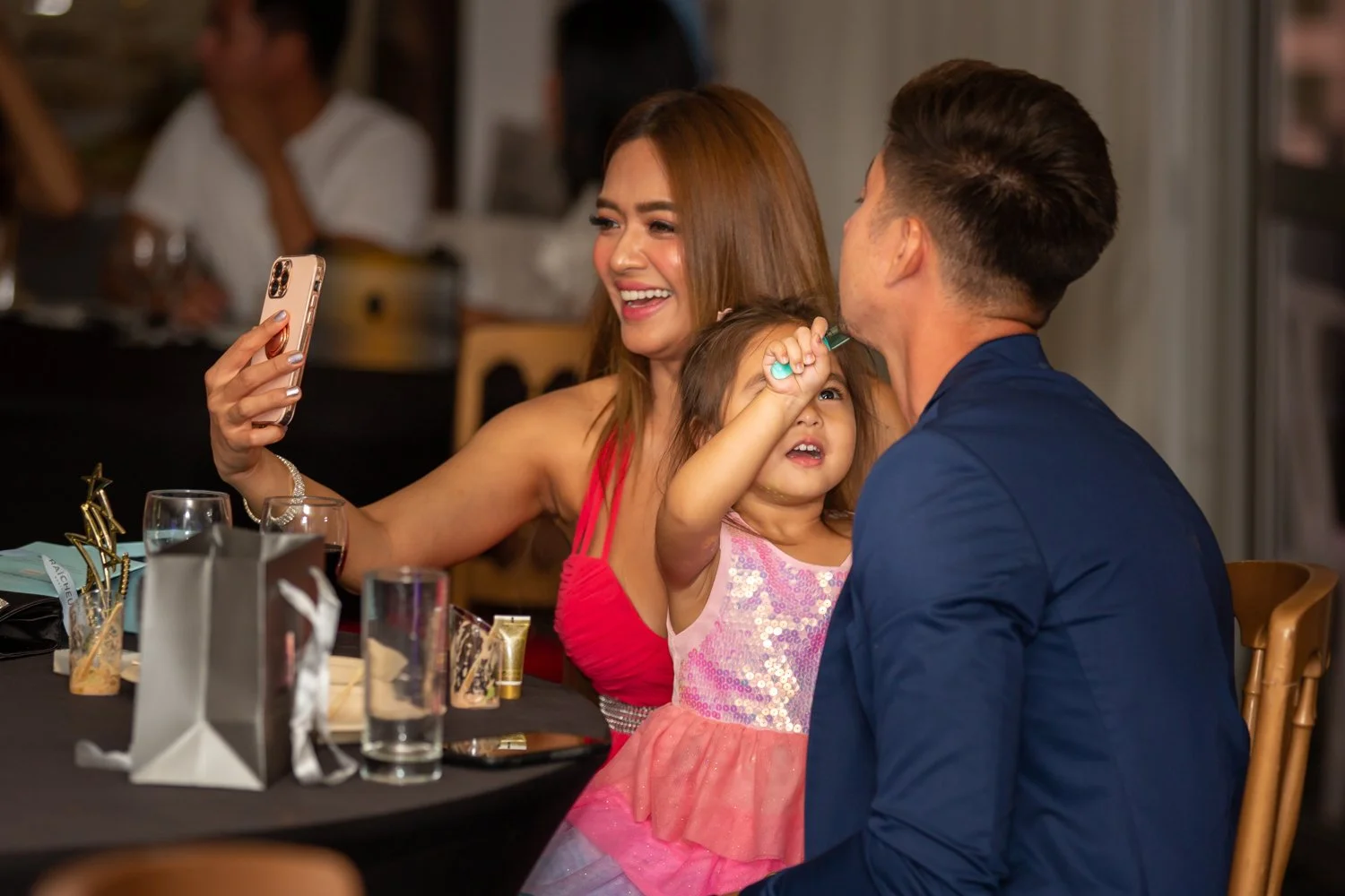 A woman and a man sitting at a dinner table with a young girl between them, who is drawing on the woman's face with a marker. The woman is smiling while taking a selfie with her phone, and the young girl looks focused.