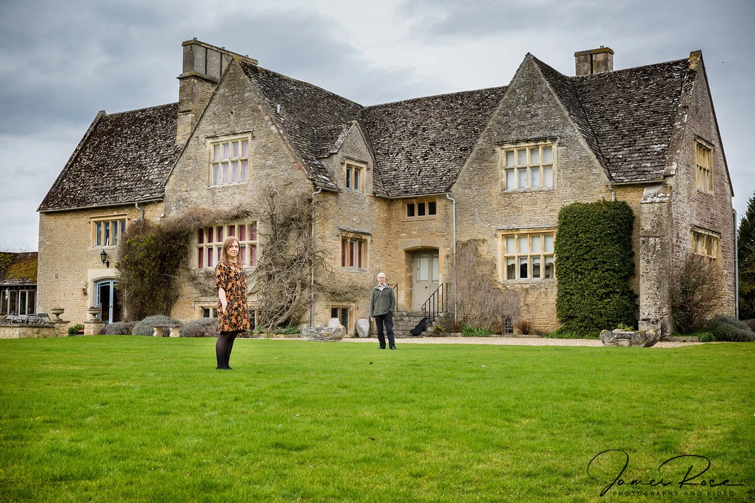 A large, old stone house with a steep, shingled roof, multiple windows, and a central front door. There are two people standing on a well-manicured lawn in front of it, one woman in a floral dress and one man in a green jacket with a black bag nearby