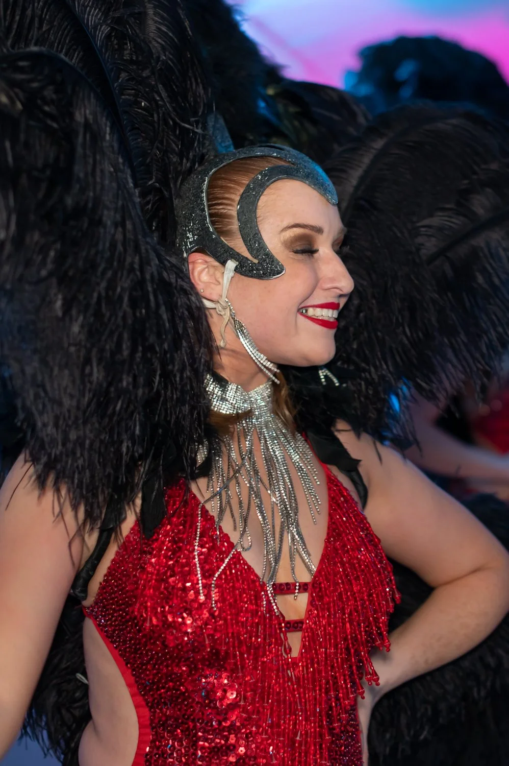 Woman in a red sequined dress with layered silver jewelry, wearing a large black feathered costume with a dramatic headpiece and face makeup, smiling with her eyes closed.
