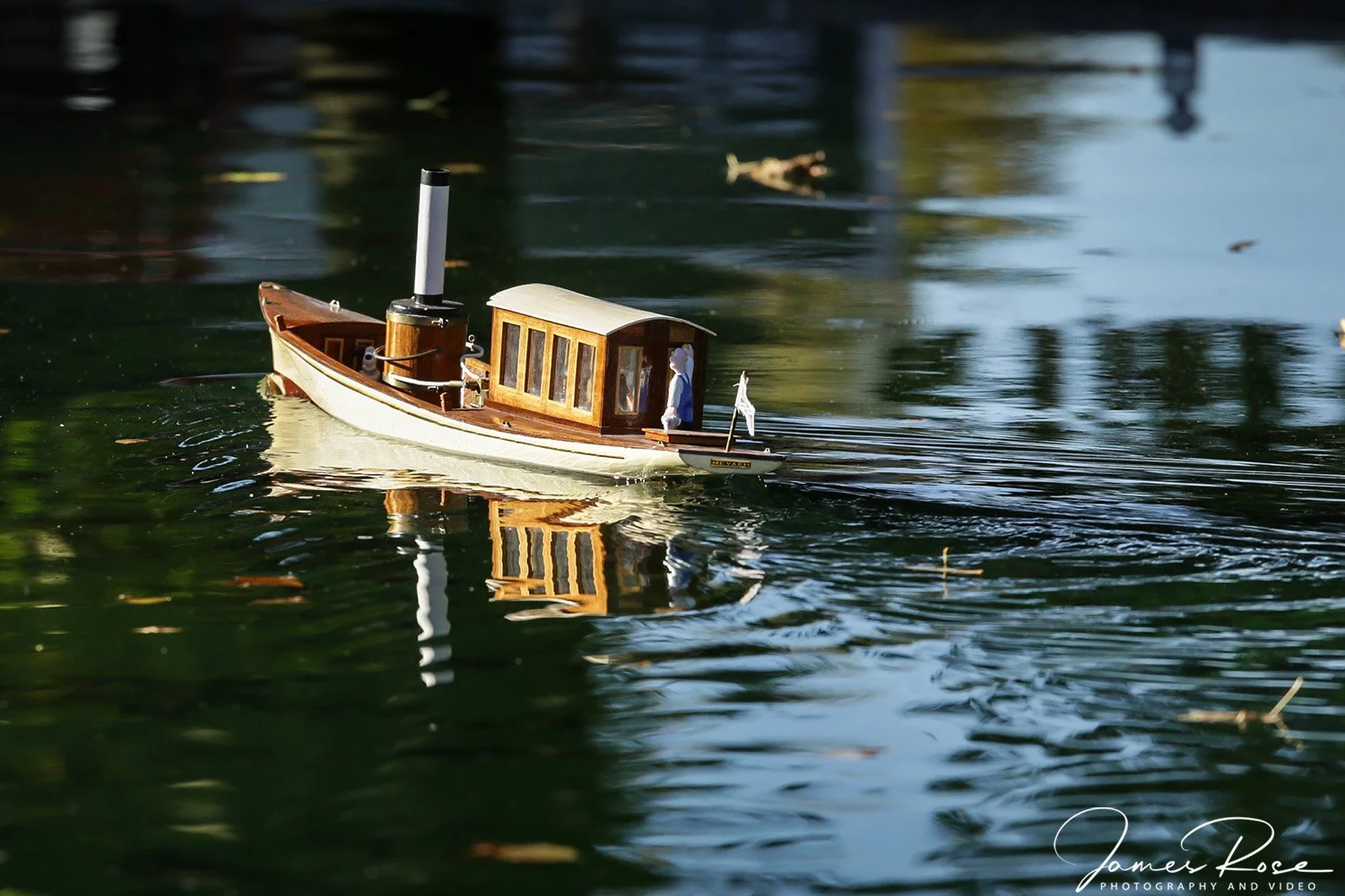 A small model boat floating on a calm body of water, reflecting the boat and surrounding trees.