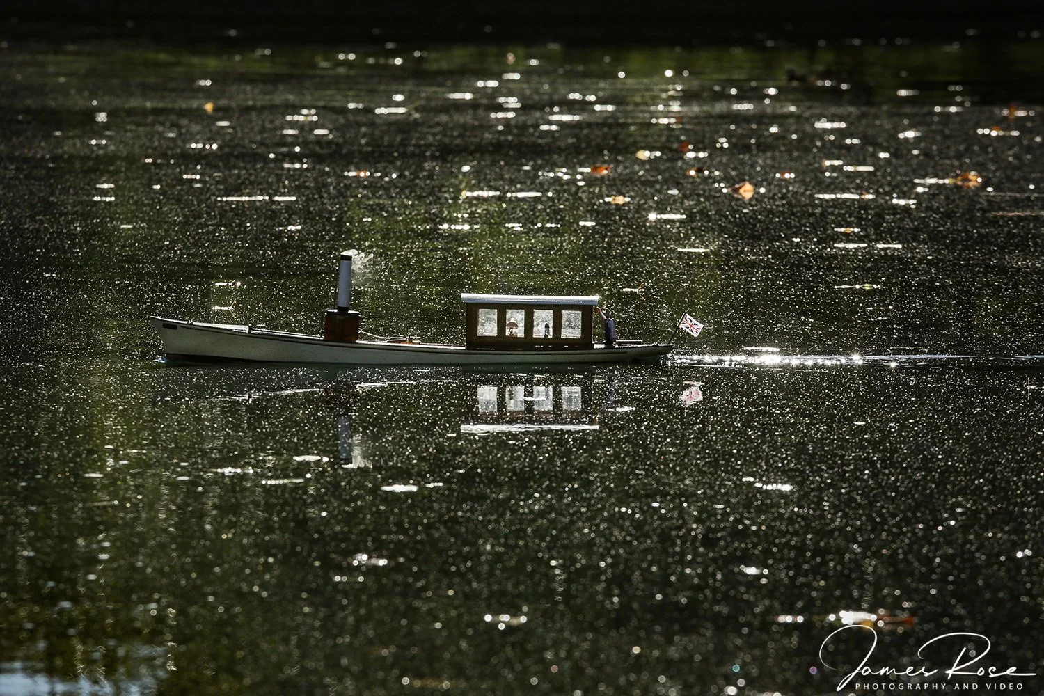 A small toy boat floating on a reflective water surface. The water is dark with glittering specks, and the boat has a small Union Jack flag at the stern.