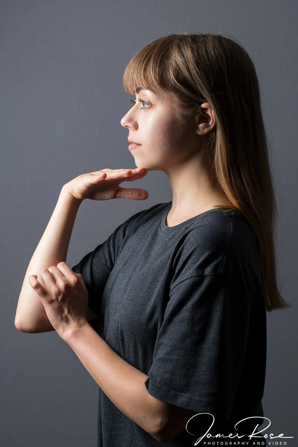 Side profile of a young woman with light brown hair, wearing a black t-shirt, posing against a plain gray background.