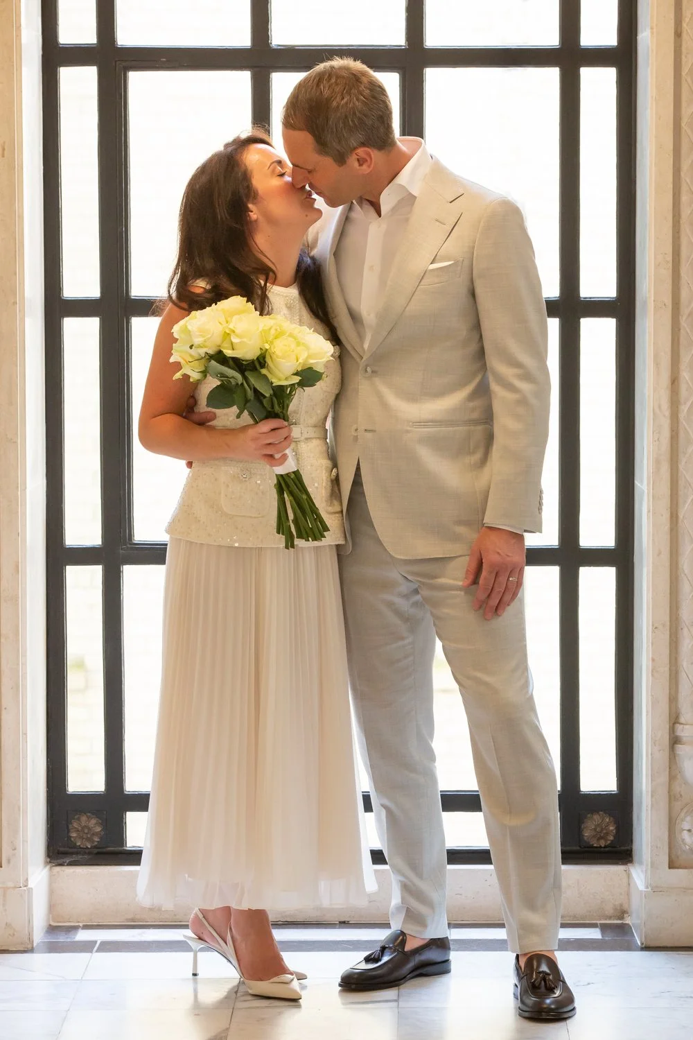 A bride and groom share an intimate moment, about to kiss, in front of a large window. The bride holds a bouquet of white roses, wearing a cream dress with a pleated skirt. The groom is dressed in a light-colored suit with a white shirt and black sho