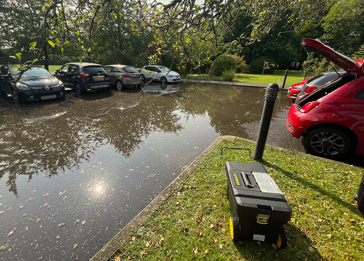 A parking lot with water accumulation after heavy rain, reflecting trees and cars, with a car's trunk open on the right. In the foreground, a portable generator is placed on the grassy area next to the parking lot.