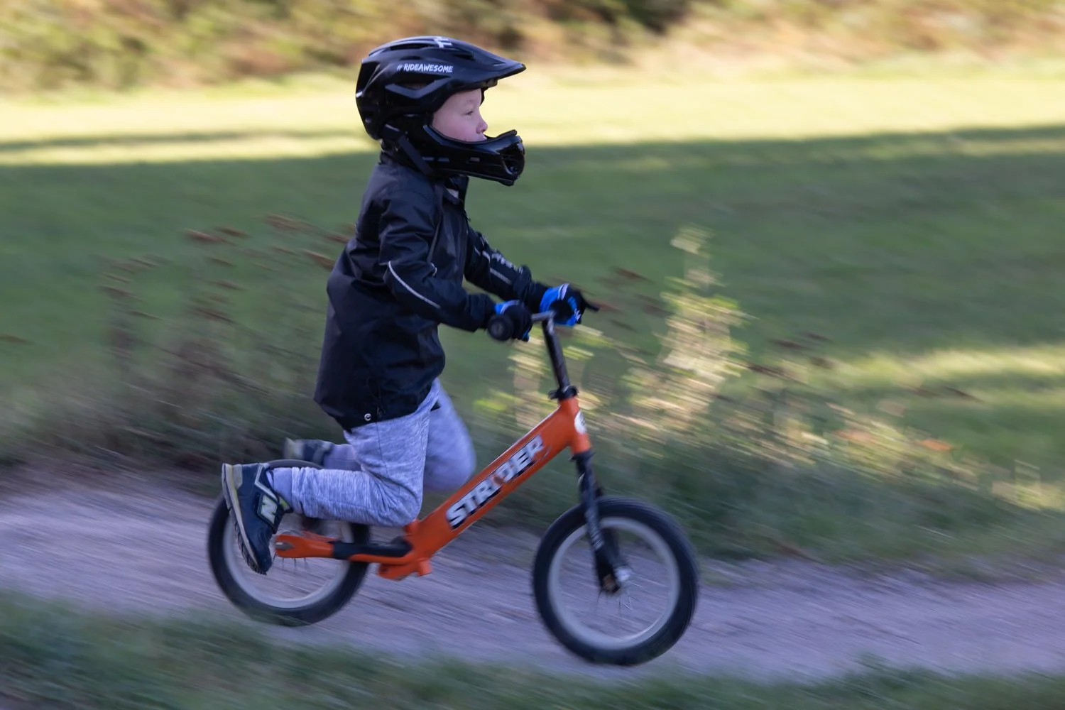 A young boy wearing a black helmet, black jacket, and gray pants riding an orange Strider balance bike on a dirt path in a grassy area.