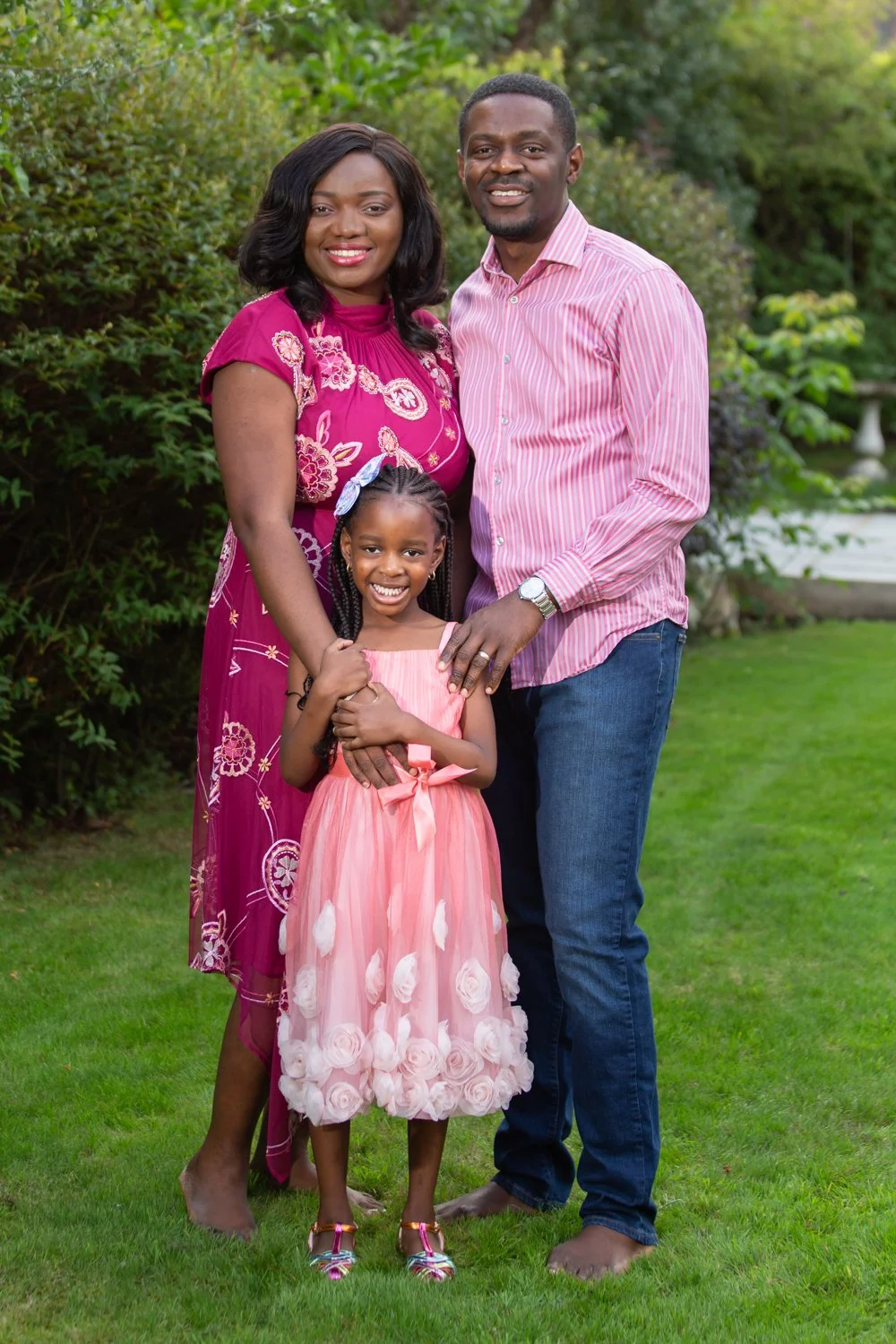 A happy family of three posing outdoors in a garden, with a woman in a pink dress, a man in a striped pink shirt, and a young girl in a pink dress with 3D floral details.