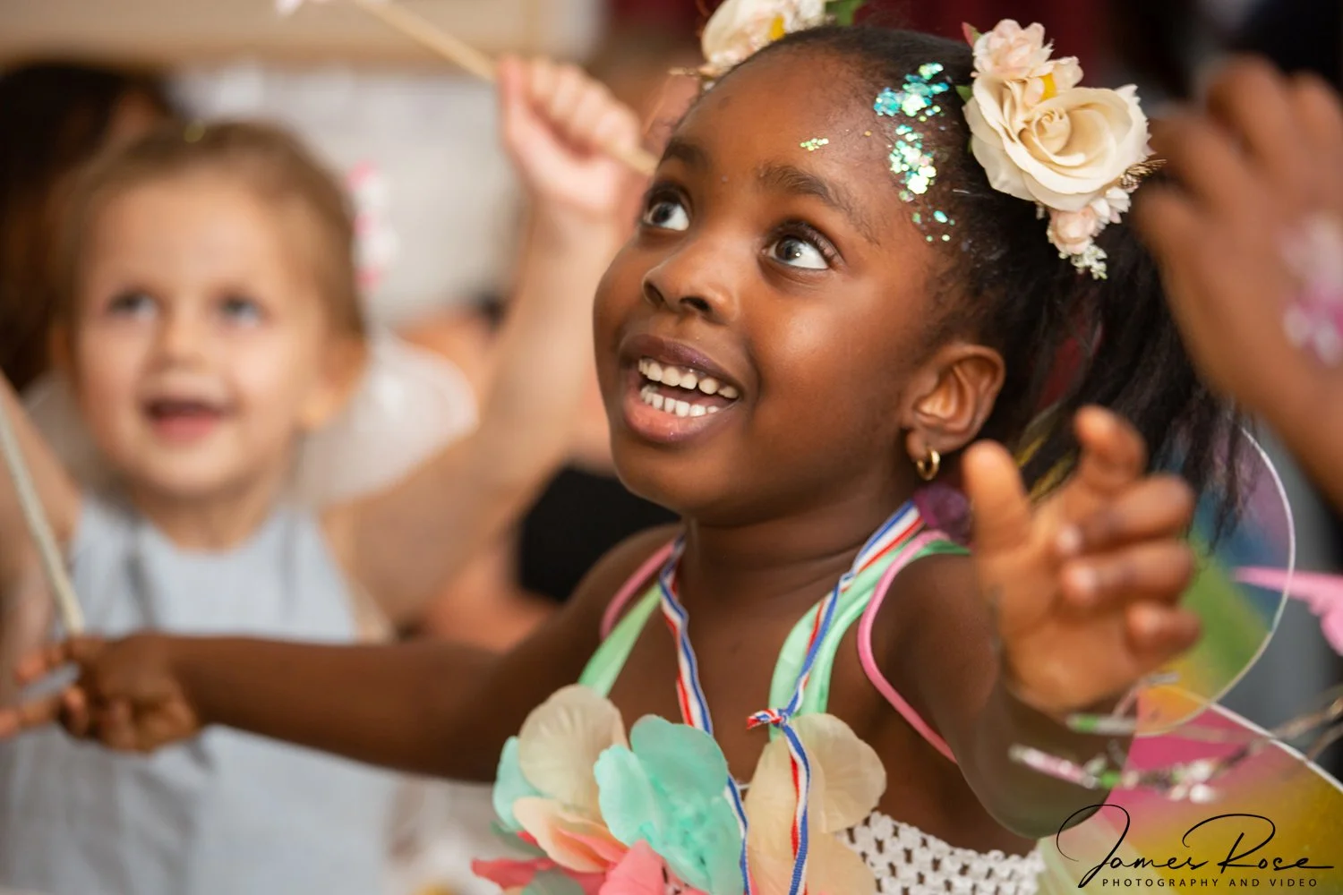 A young girl with floral headband and colorful lei celebrating joyfully with other children at a party or gathering.